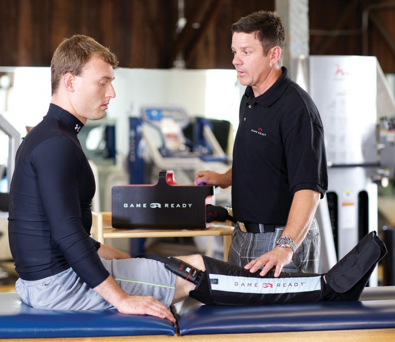A physiotherapist fitting a Game Ready leg wrap on an athlete in a rehab gym, with the GRPro 2.1 unit on the plinth beside them