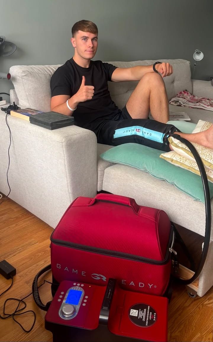 Meath footballer Ruairí Kinsella on his sofa at home giving a thumbs up, wearing a Game Ready leg wrap with the Game Ready unit beside him