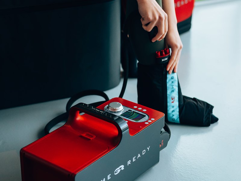 A woman fitting a Game Ready ankle wrap, with the GRPro 2.1 unit on the floor beside her