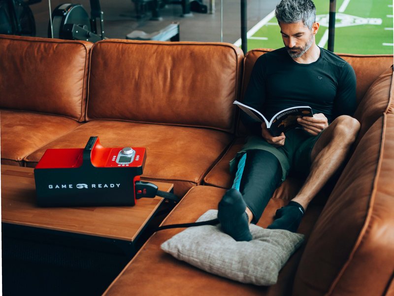 A man reading on a sofa in a training facility with a Game Ready wrap on his leg and the Game Ready unit beside him