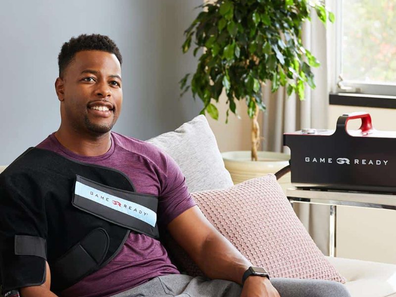 A man smiling on his sofa at home wearing the Game Ready shoulder wrap, with the Game Ready unit on a side table